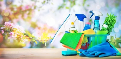 Assortment of household cleaning products including spray bottles, gloves, microfiber cloths, sponges, and a squeegee arranged on a wooden table