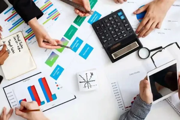 Top-down view of a business meeting with several people reviewing charts and financial documents on a table. Hands are seen using calculators, smartphones, a tablet, and writing notes while discussing colorful graphs and data reports.