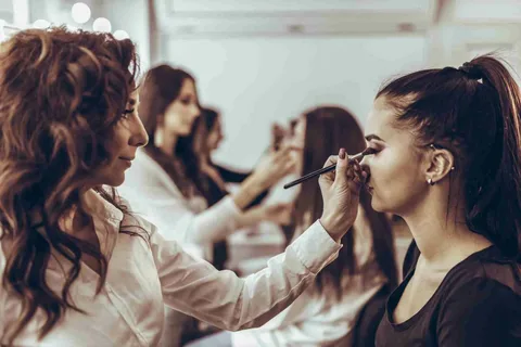 A girl applying makeup while looking into a mirror, with cosmetic products such as eyeshadow palette, foundation bottle, makeup brushes, and lipstick arranged on a table in front of her