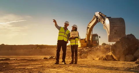 Exterior view of a construction company site or office, with company signage, construction vehicles (such as excavators, dump trucks, or cranes), building materials (pipes, lumber, or gravel piles), portable trailers, and workers in hard hats and high-visibility vests