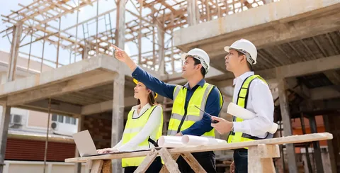 Construction company workers and heavy machinery erecting a building structure, with steel framework, concrete foundations, cranes lifting materials, scaffolding around partially completed floors, and workers in hard hats and safety vests on site.