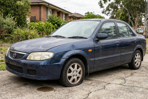 A damaged, unwanted junk car with rust spots and a flat tire sitting unused on a residential driveway.